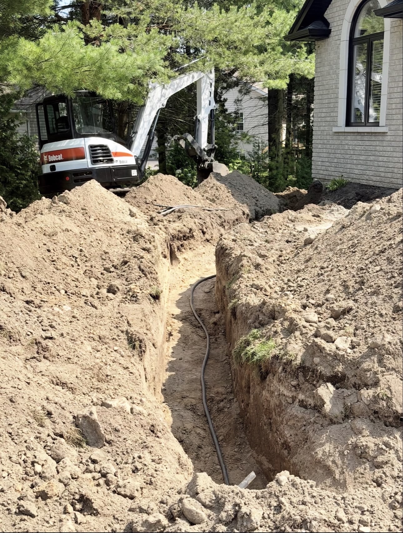 Open excavated trench with black drainage pipe laid in the bottom running toward a house