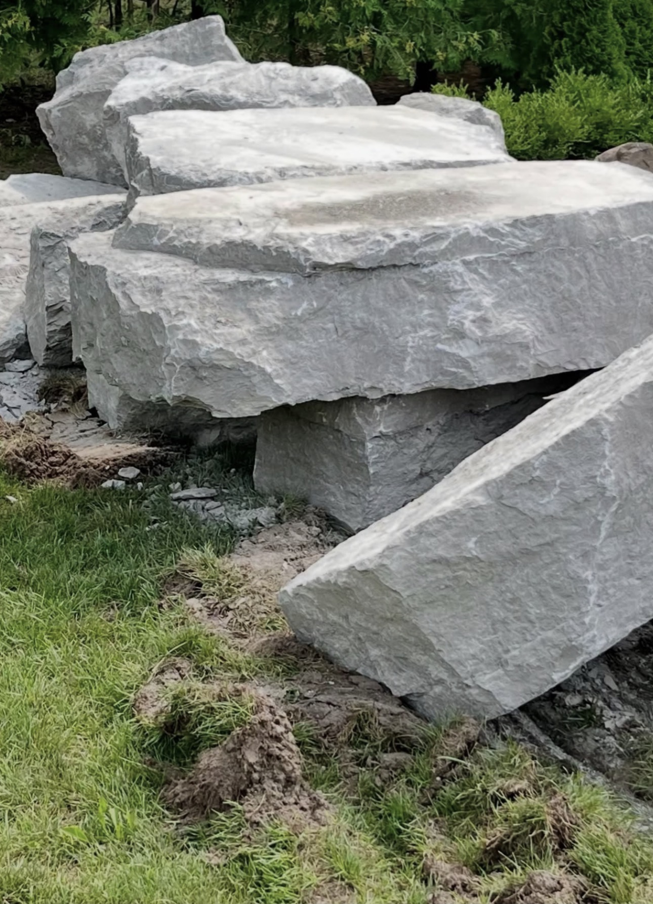 Large flat armour stone slabs stacked on a lawn beside cedar trees