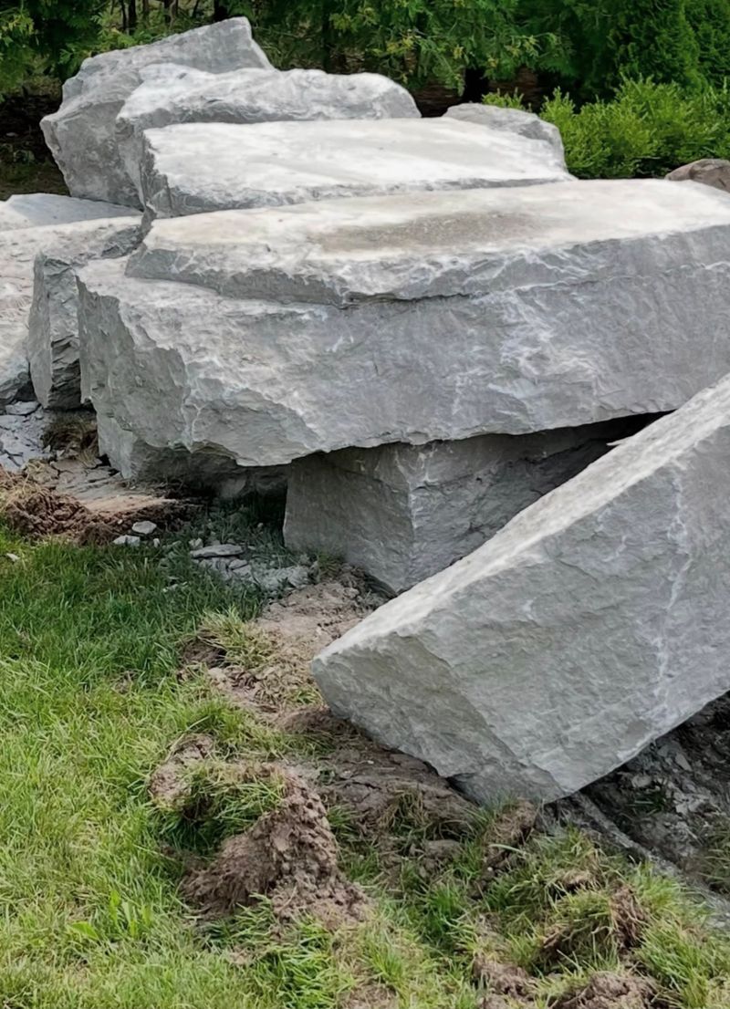 Large flat armour stone slabs stacked on a lawn beside cedar trees