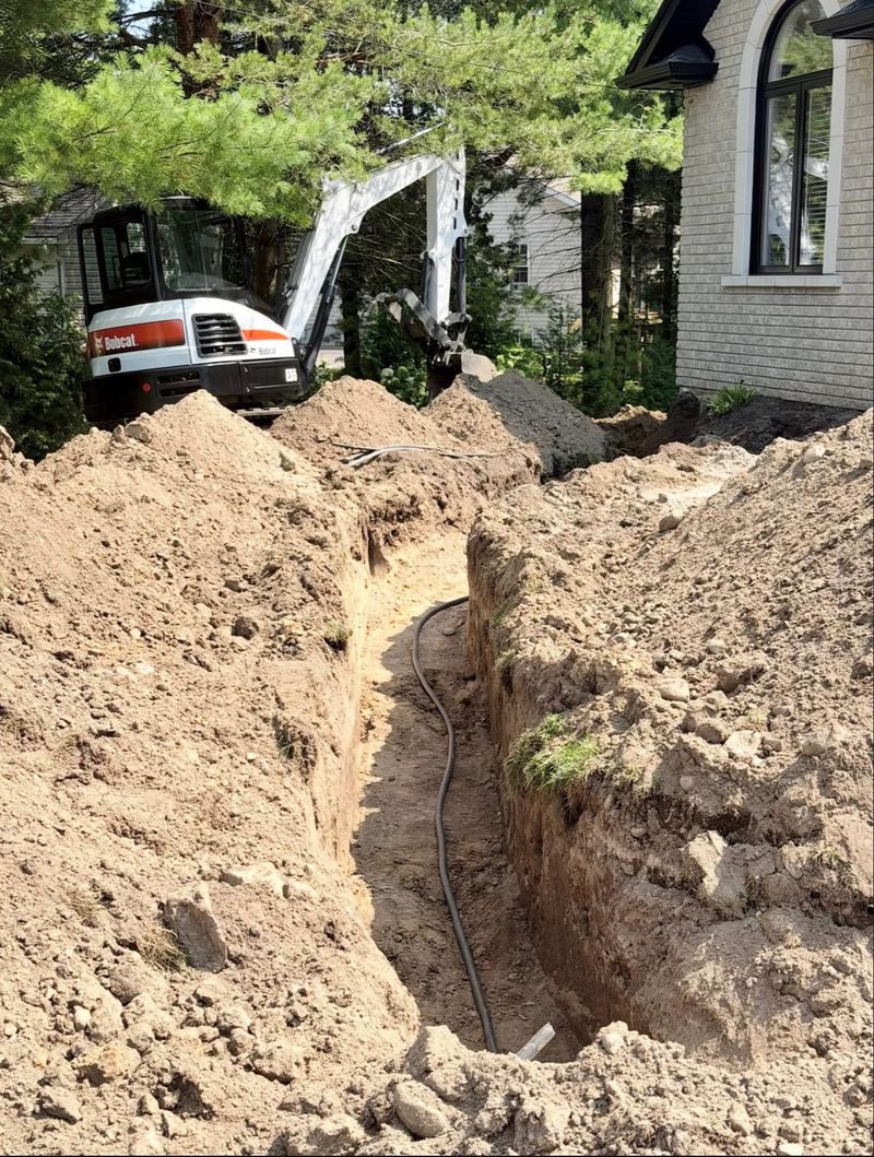 Open excavated trench with black drainage pipe laid in the bottom running toward a house