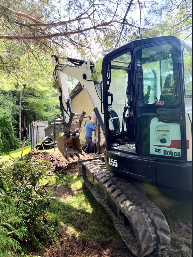 Bobcat E55 mini excavator digging a trench alongside a brick house while a worker checks the line